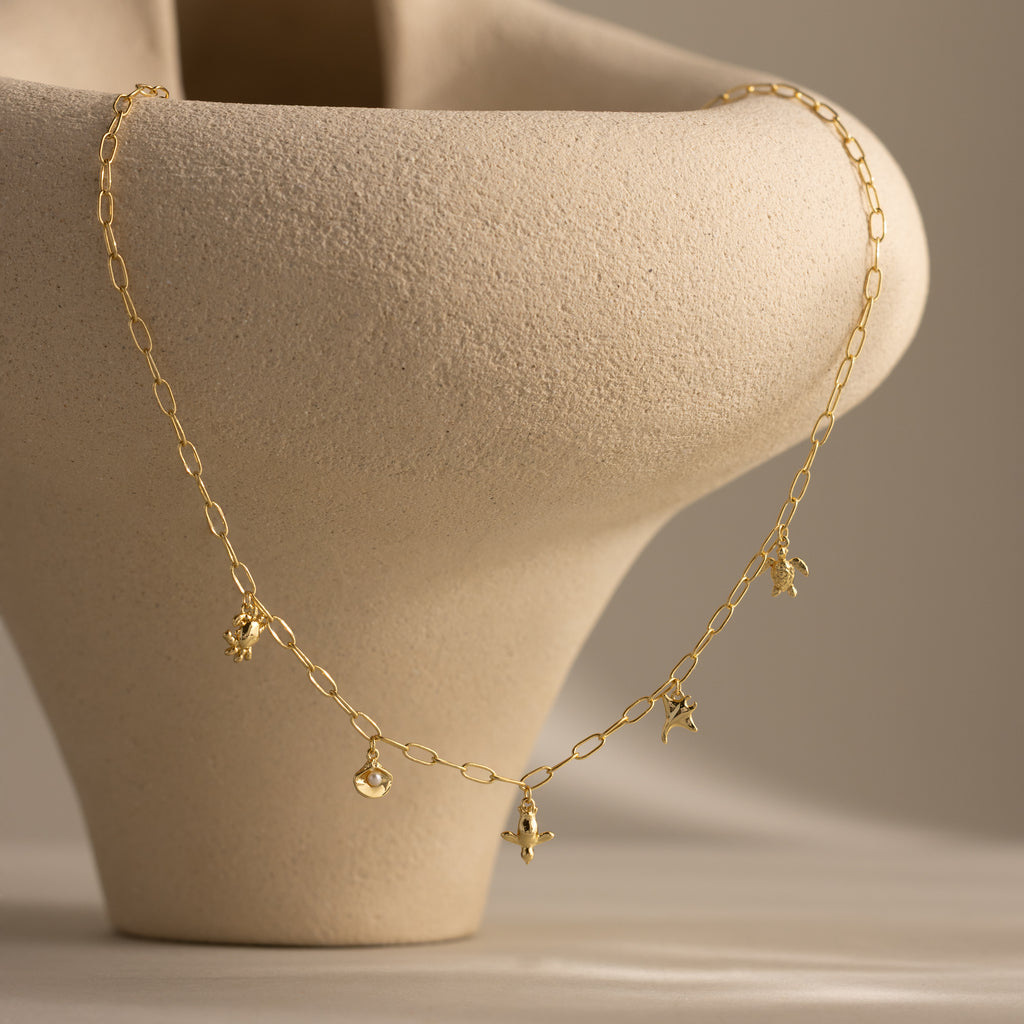 Seaside Charm Necklace in 18K Gold features delicate star and round charms, elegantly draped over a textured beige ceramic vase—an ideal piece of beach-inspired jewelry.