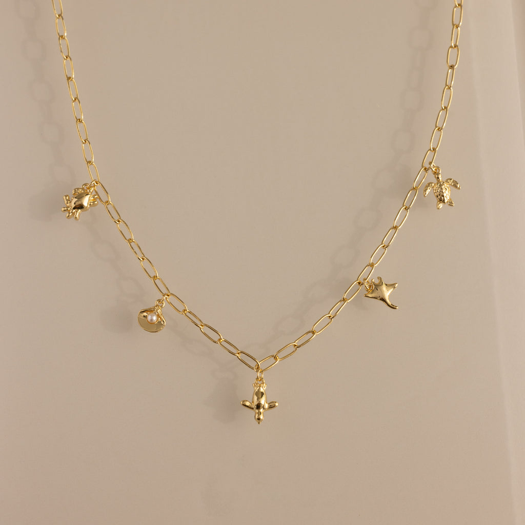 The Seaside Charm Necklace in 18K Gold features five delicate charms—a bird, leaf, flower, and round pendant—perfect beach jewelry for summer lovers of dainty style.
