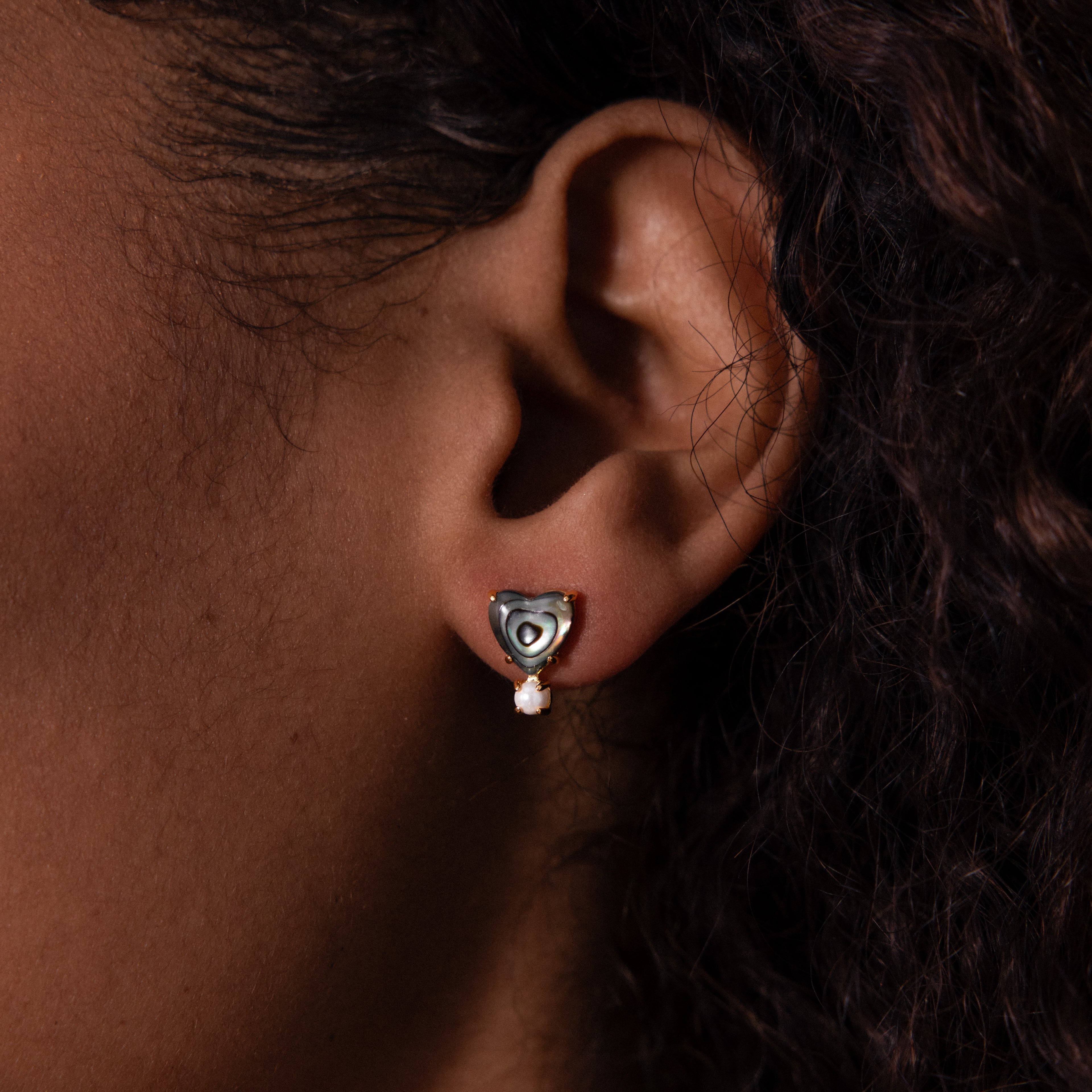 A close-up of an ear wearing Abalone Pearl Heart Studs—heart-shaped silver studs with a dangling pearl—captures the delicate beauty of coastal jewelry.