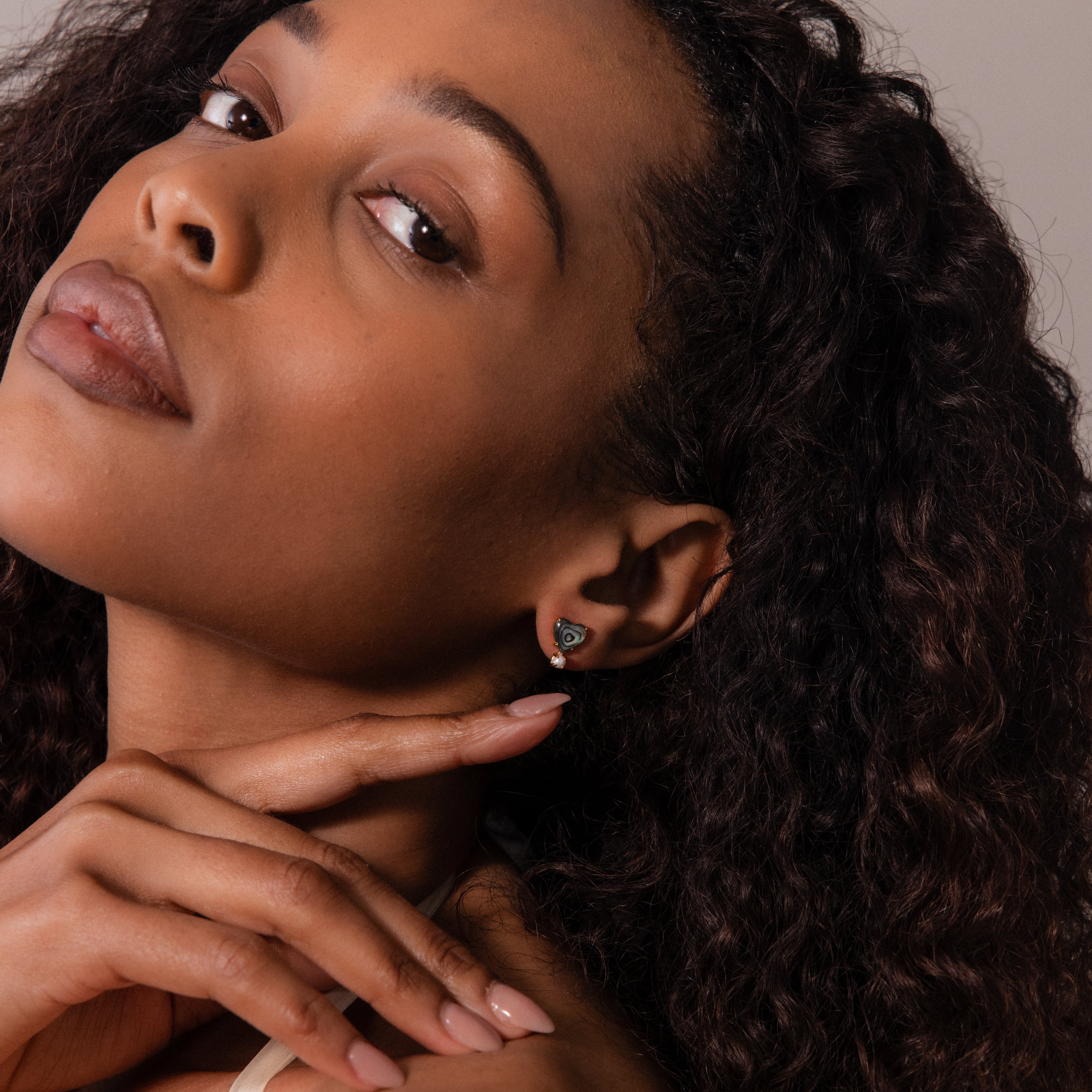 A woman with curly hair and subtle makeup glances over her shoulder, highlighting her manicured nails and the Abalone Pearl Heart Studs—a chic piece of coastal-inspired jewelry.