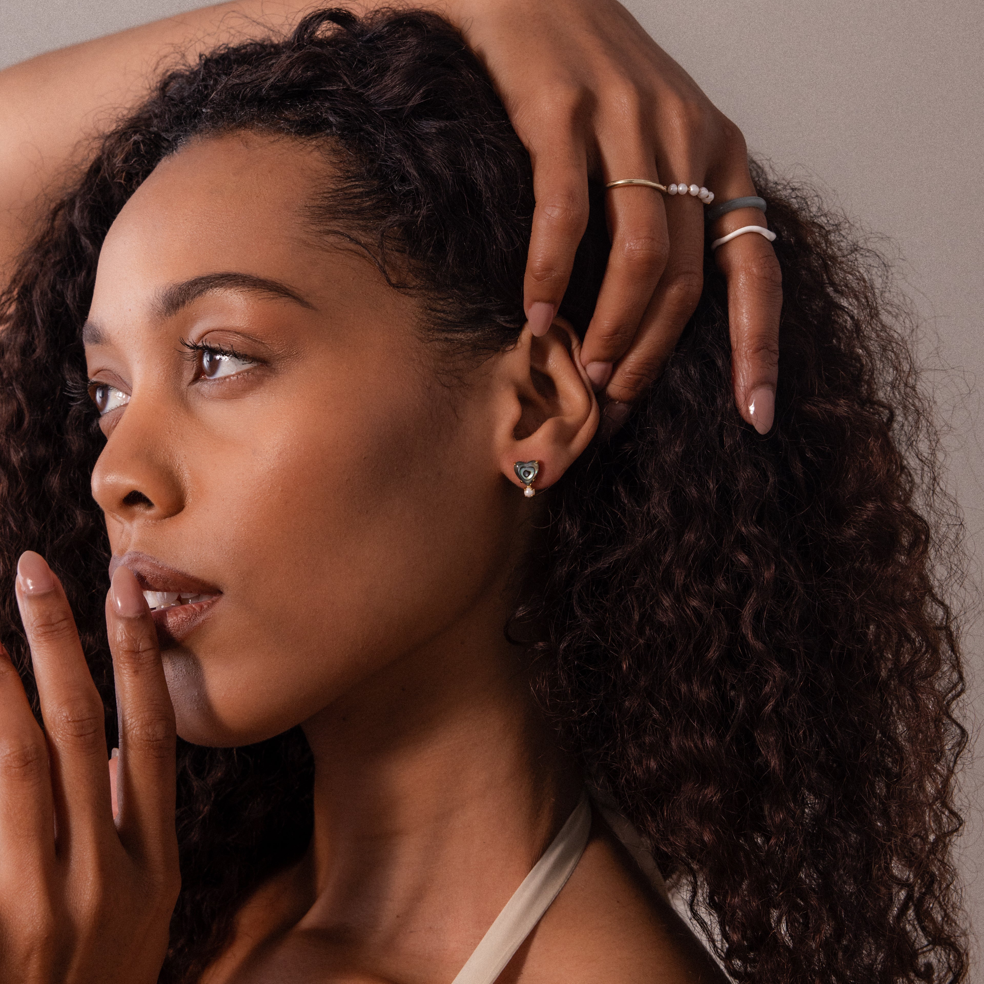 A woman with wavy hair touches her face, wearing pearl rings and Abalone Pearl Heart Studs, looking thoughtful in elegant coastal jewelry by Abalone Pearl Heart Studs.