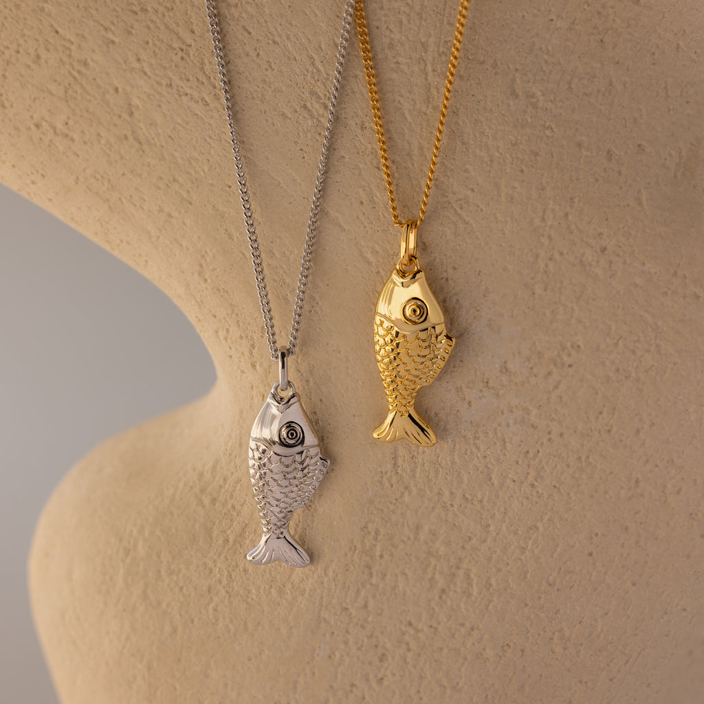 The Fish Pendant Necklace features silver and gold fish-shaped pendants on chains, displayed on a textured beige background—an ocean-inspired piece ideal for layering.