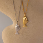 The Fish Pendant Necklace features silver and gold fish-shaped pendants on chains, displayed on a textured beige background—an ocean-inspired piece ideal for layering.