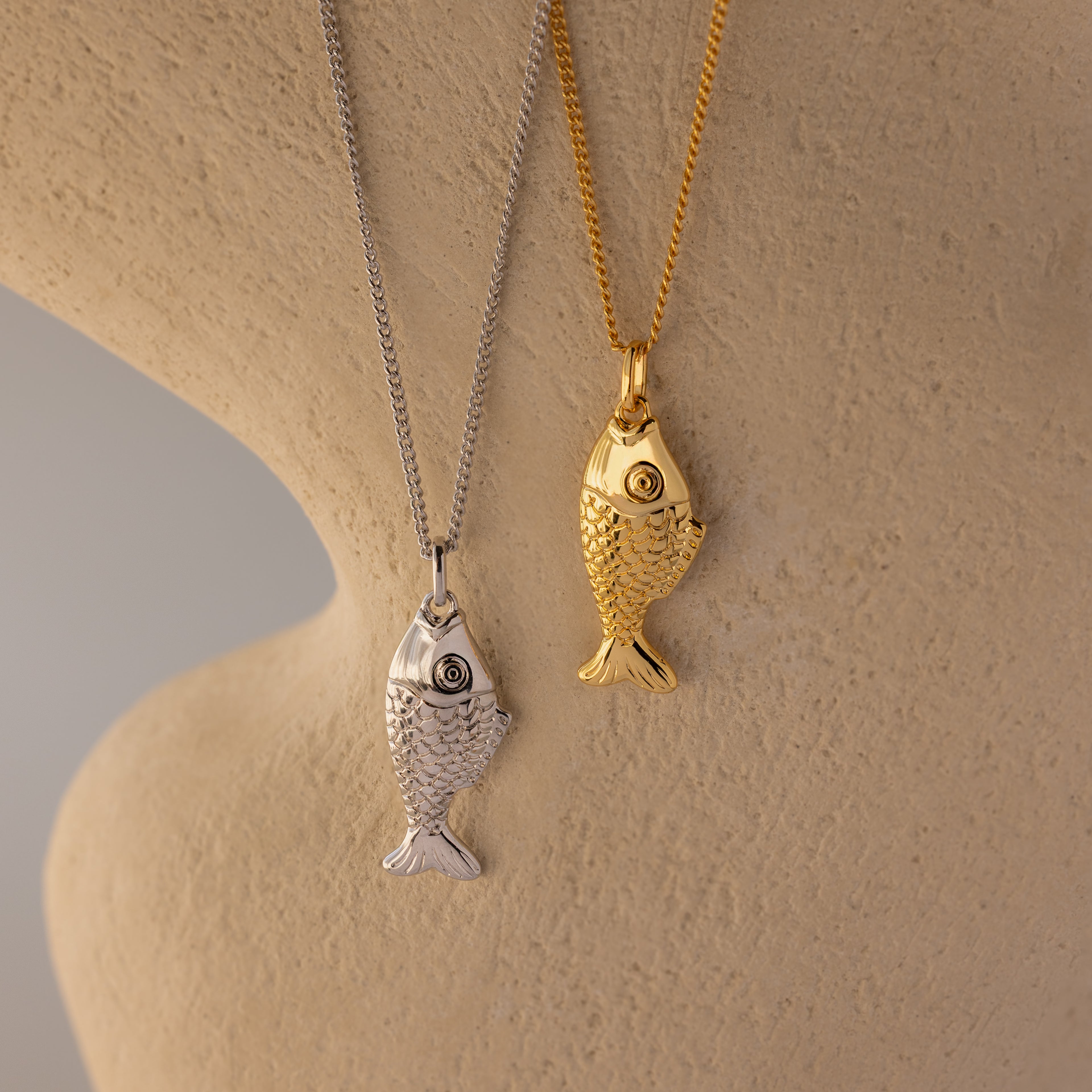 The Fish Pendant Necklace features silver and gold fish-shaped pendants on chains, displayed on a textured beige background—an ocean-inspired piece ideal for layering.