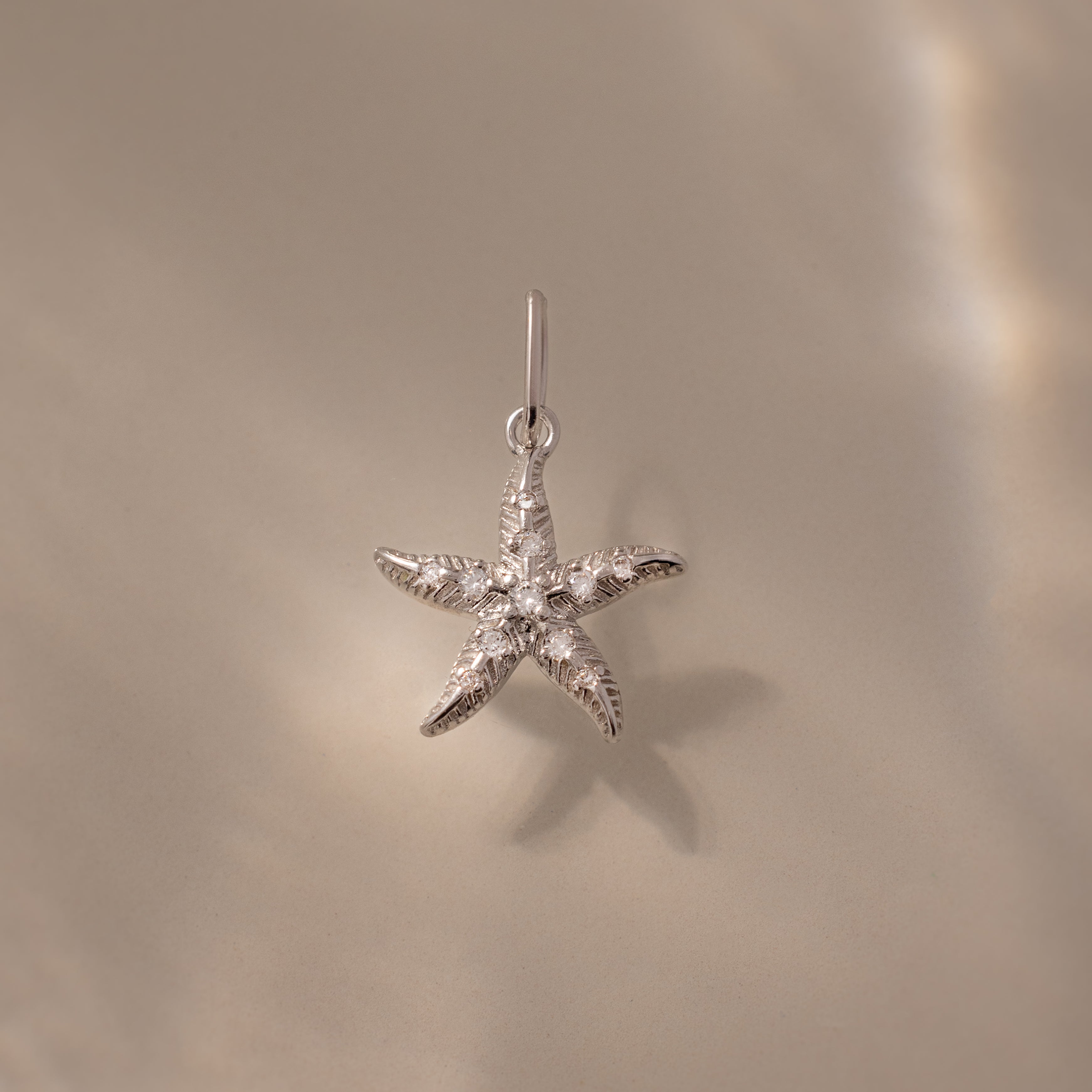 The Pave Starfish Charm in Silver is displayed on a smooth beige background with soft light reflections, highlighting the elegant coastal charm of this starfish jewelry.