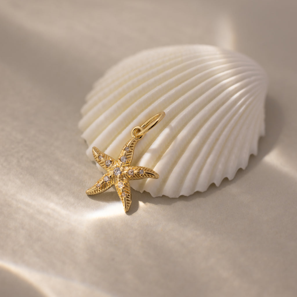 The Pave Starfish Charm in 18K Gold, adorned with small gems for coastal flair, is displayed beside a white seashell on soft sand.