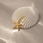 The Pave Starfish Charm in 18K Gold, adorned with small gems for coastal flair, is displayed beside a white seashell on soft sand.