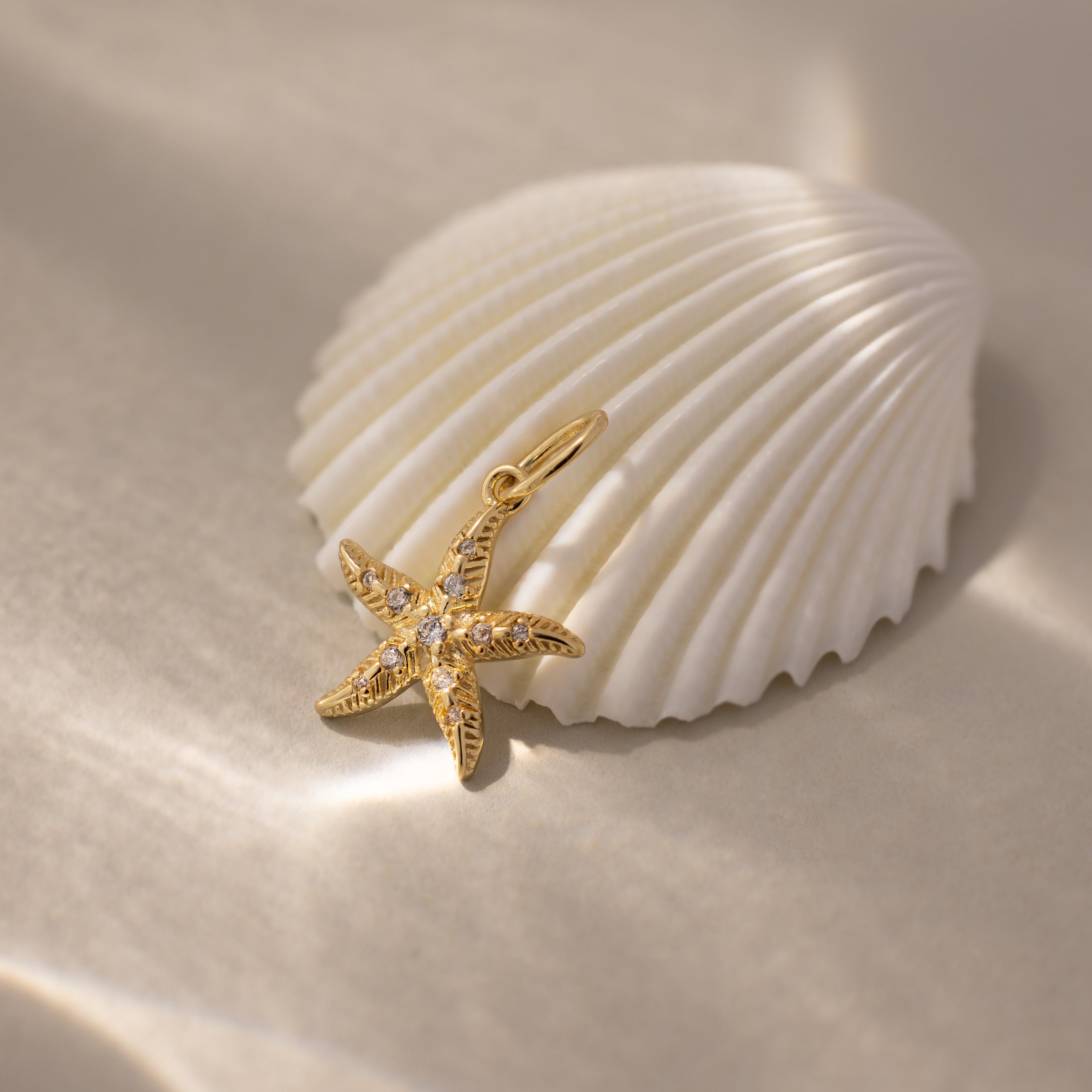 The Pave Starfish Charm in 18K Gold, adorned with small gems for coastal flair, is displayed beside a white seashell on soft sand.