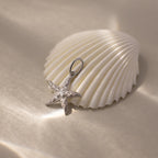 The Pave Starfish Charm in Silver displayed on a white seashell with soft light and shadows, evoking timeless coastal charm against a beige background.