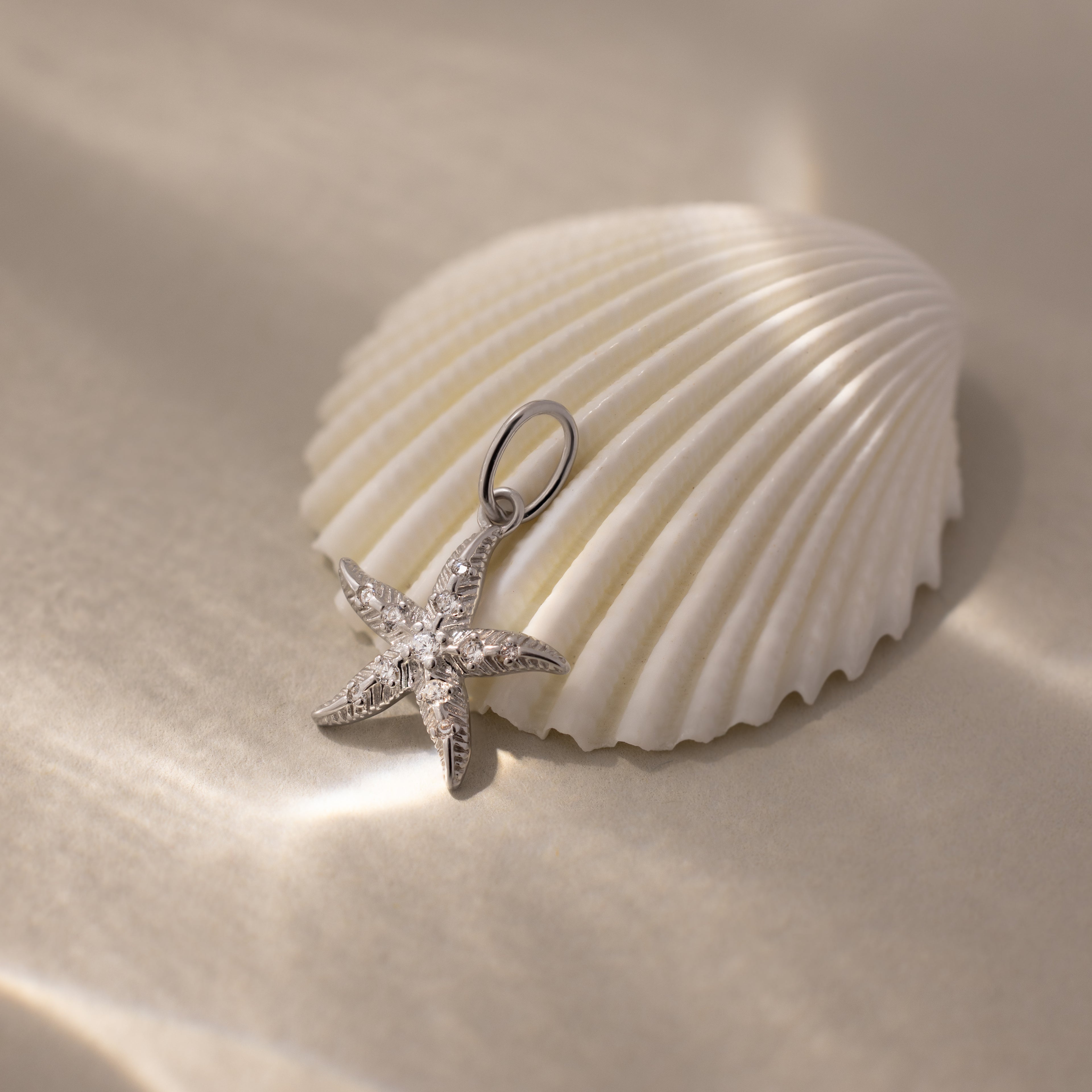 The Pave Starfish Charm in Silver displayed on a white seashell with soft light and shadows, evoking timeless coastal charm against a beige background.
