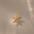 The Pave Starfish Charm in 18K Gold features shimmering gemstones, photographed on a beige surface with soft light—a stunning piece of coastal-inspired jewelry.