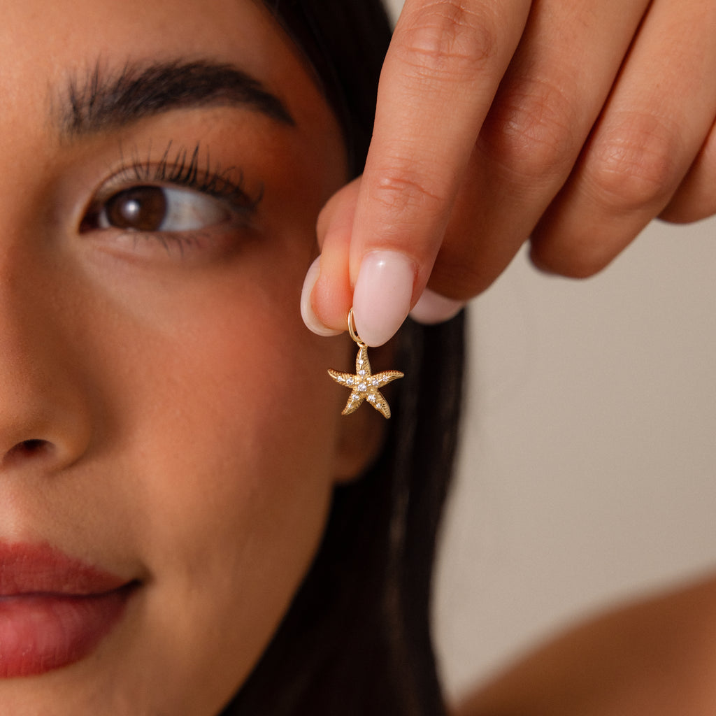 Woman holding a Pave Starfish Charm in 18K Gold close to her face, showcasing its detailed features, natural makeup, and the coastal charm of this unique gold starfish jewelry.