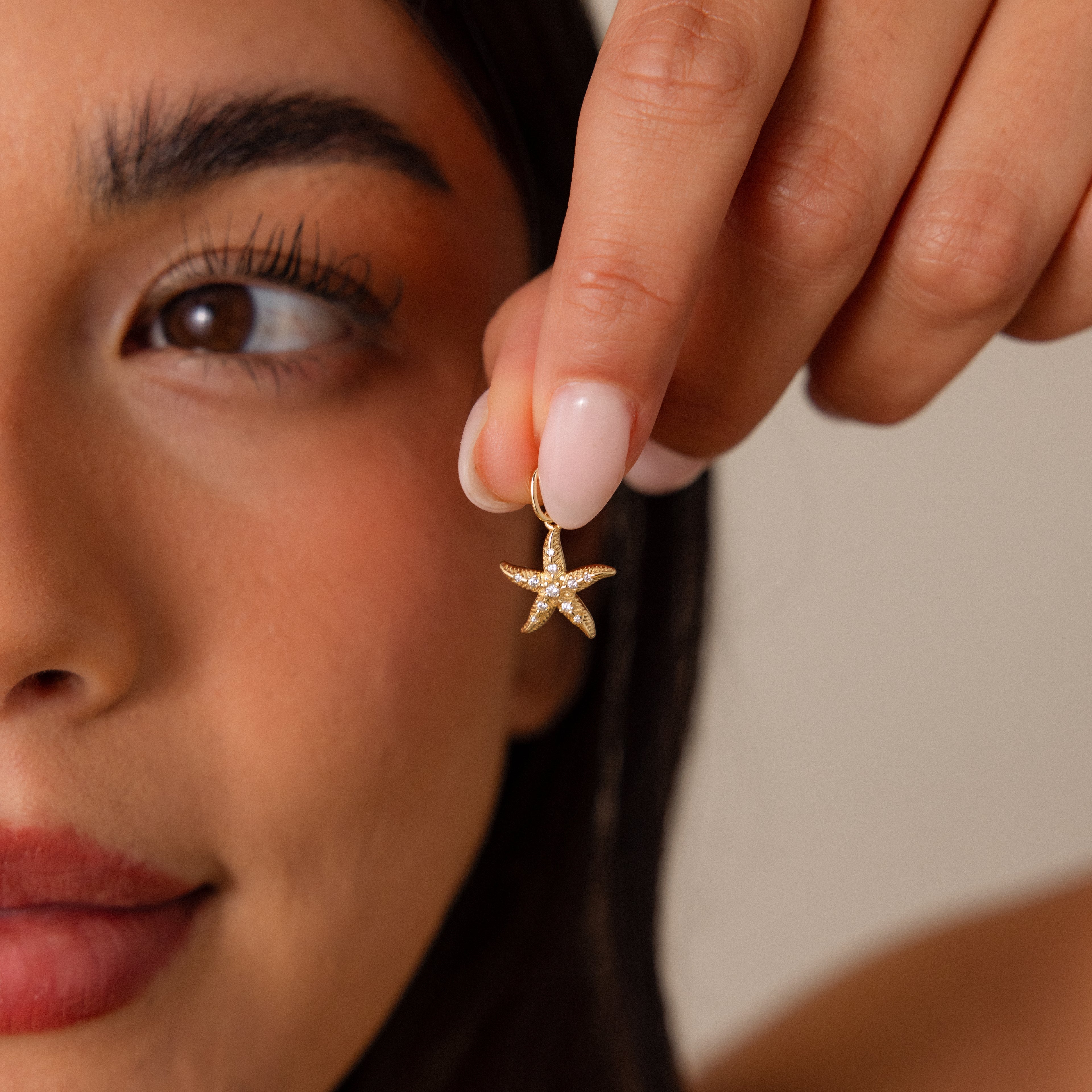 Woman holding a Pave Starfish Charm in 18K Gold close to her face, showcasing its detailed features, natural makeup, and the coastal charm of this unique gold starfish jewelry.