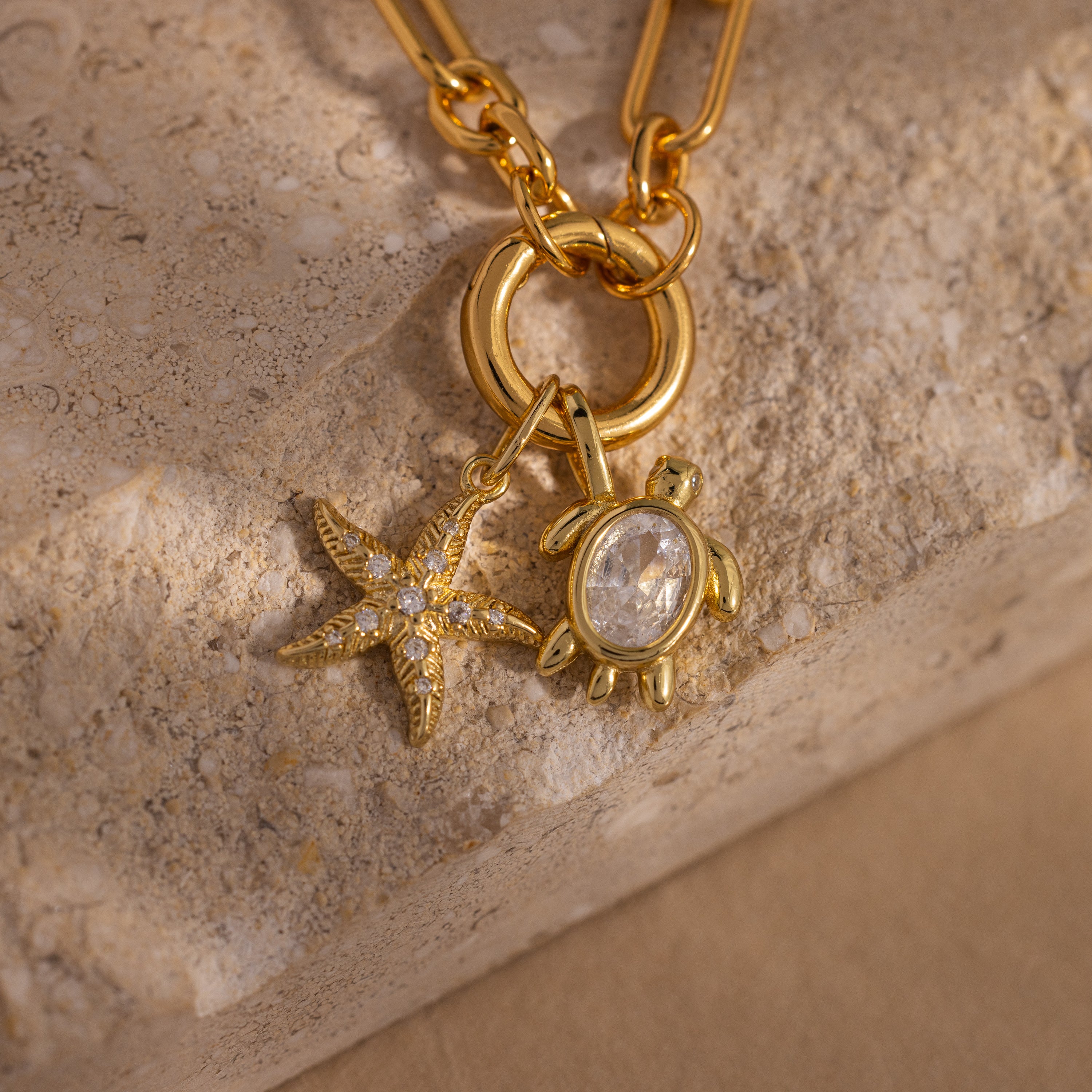 The Diamond Turtle Charm in 18K Gold, featuring clear stones and a diamond-detailed shell, creates stunning ocean-inspired jewelry when paired with a gold necklace on a textured stone surface.