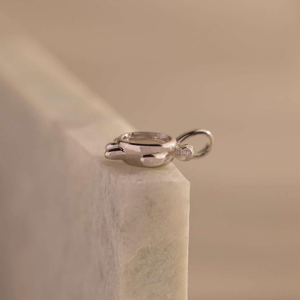 The Diamond Turtle Charm in Silver, featuring a sparkling gemstone, rests on a light marble surface—an ideal piece for admirers of ocean-inspired jewelry.