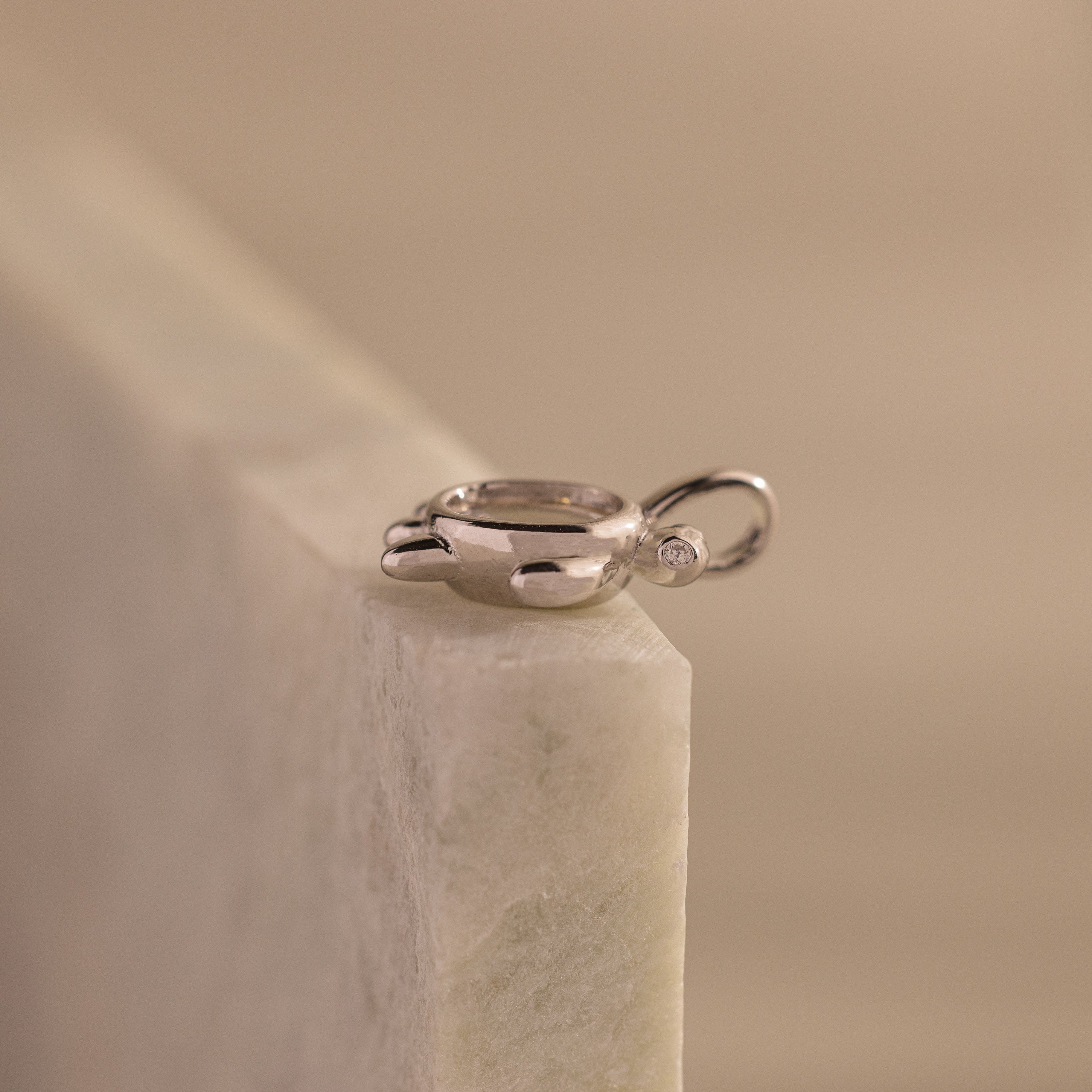 The Diamond Turtle Charm in Silver, featuring a sparkling gemstone, rests on a light marble surface—an ideal piece for admirers of ocean-inspired jewelry.
