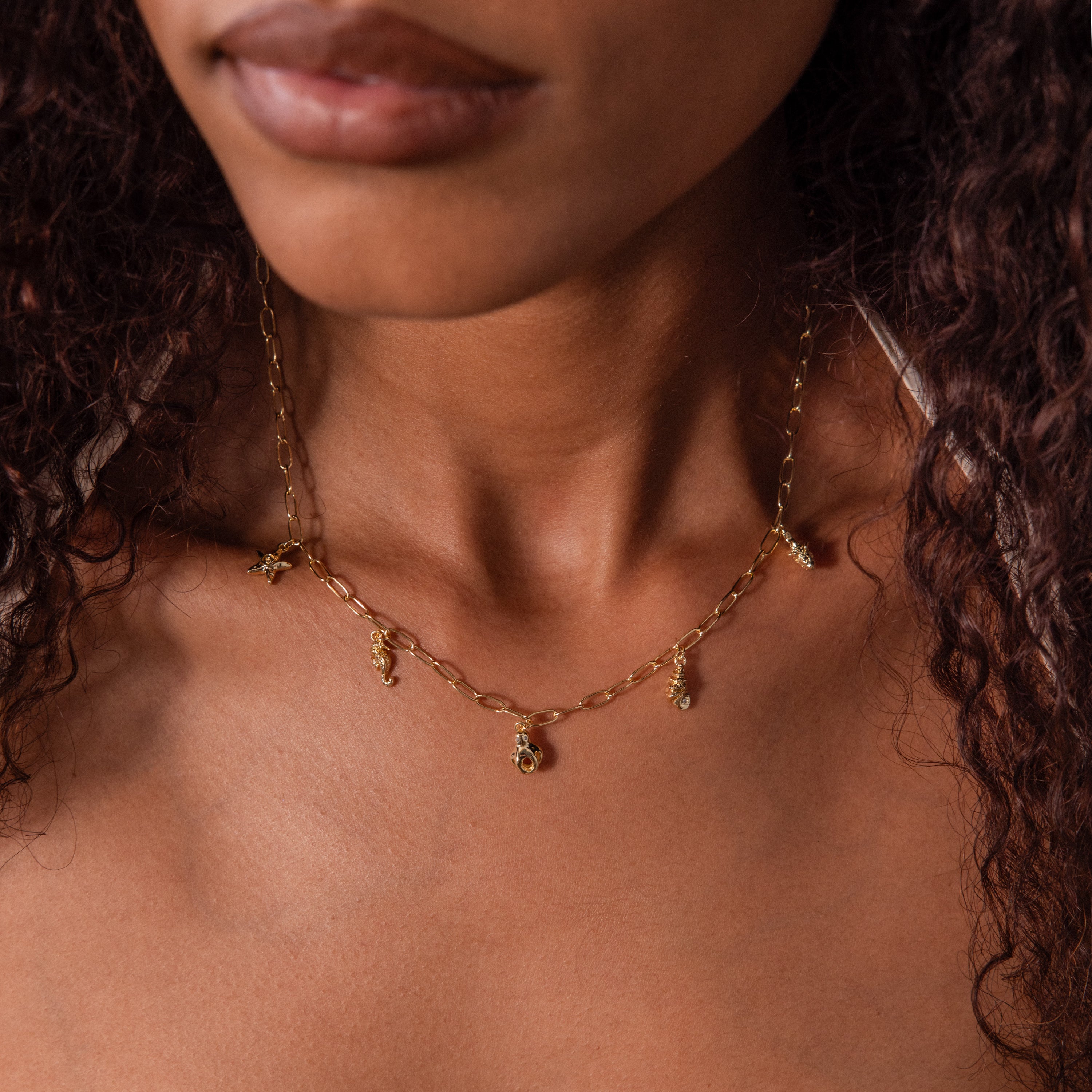 A woman wears the Ocean Charm Necklace, its delicate gold charms shown from shoulders to just below her lips, perfectly capturing seaside elegance.