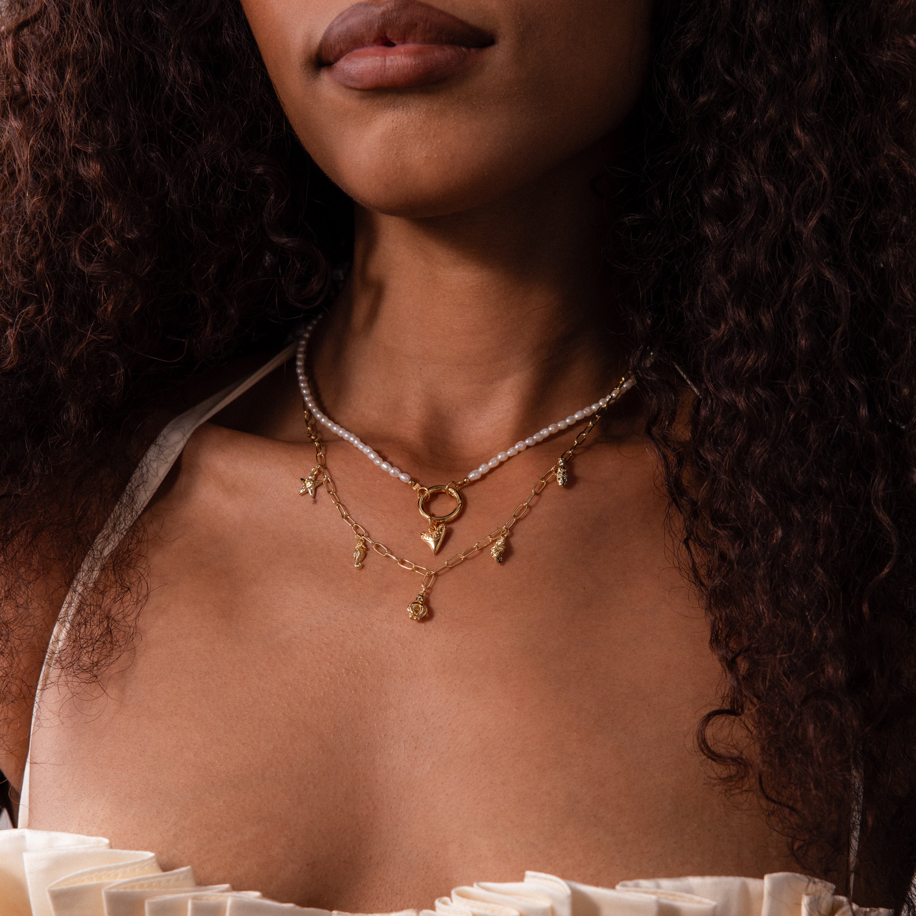 A woman shown from the shoulders up wears the Ocean Charm Necklace layered with gold necklaces and a cream ruffled top.