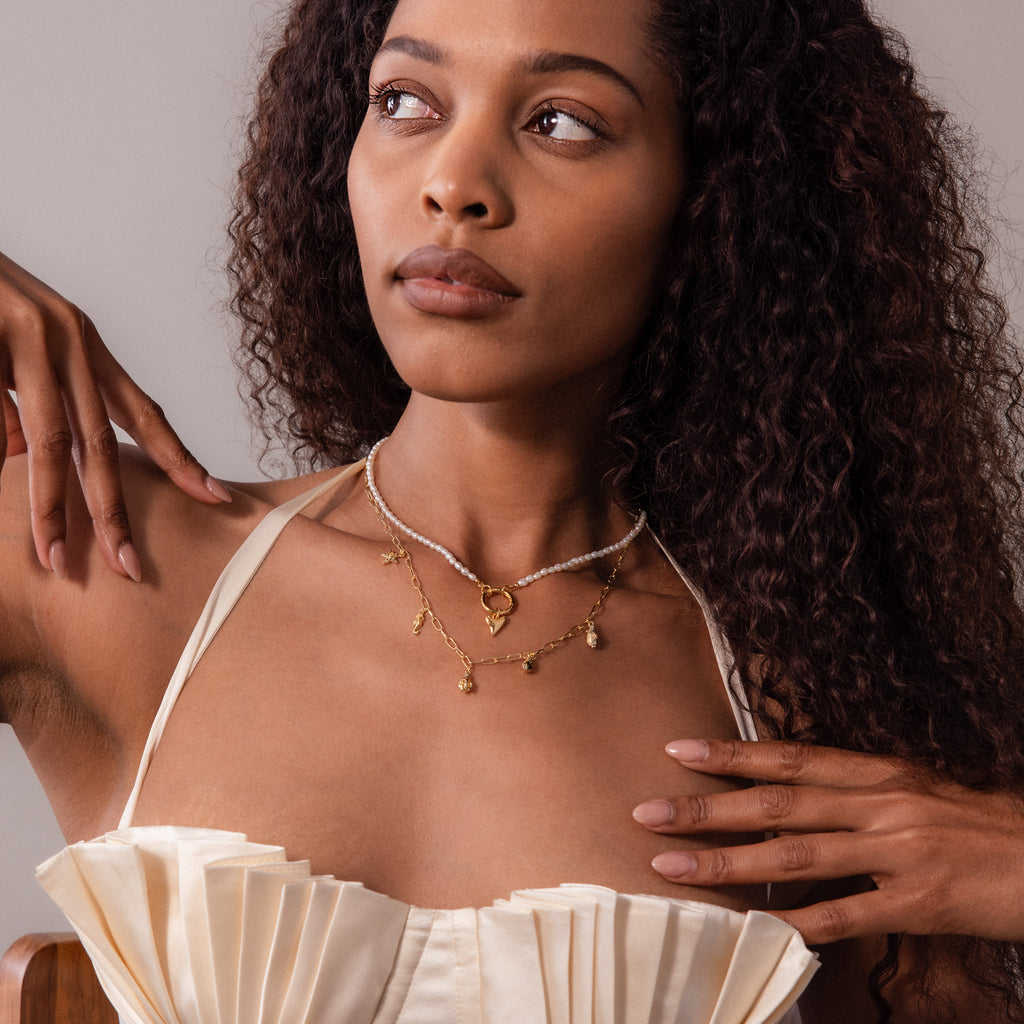 Woman with long curly hair wearing a cream pleated top and layered gold necklaces, featuring the Ocean Charm Necklace, looking to the side.