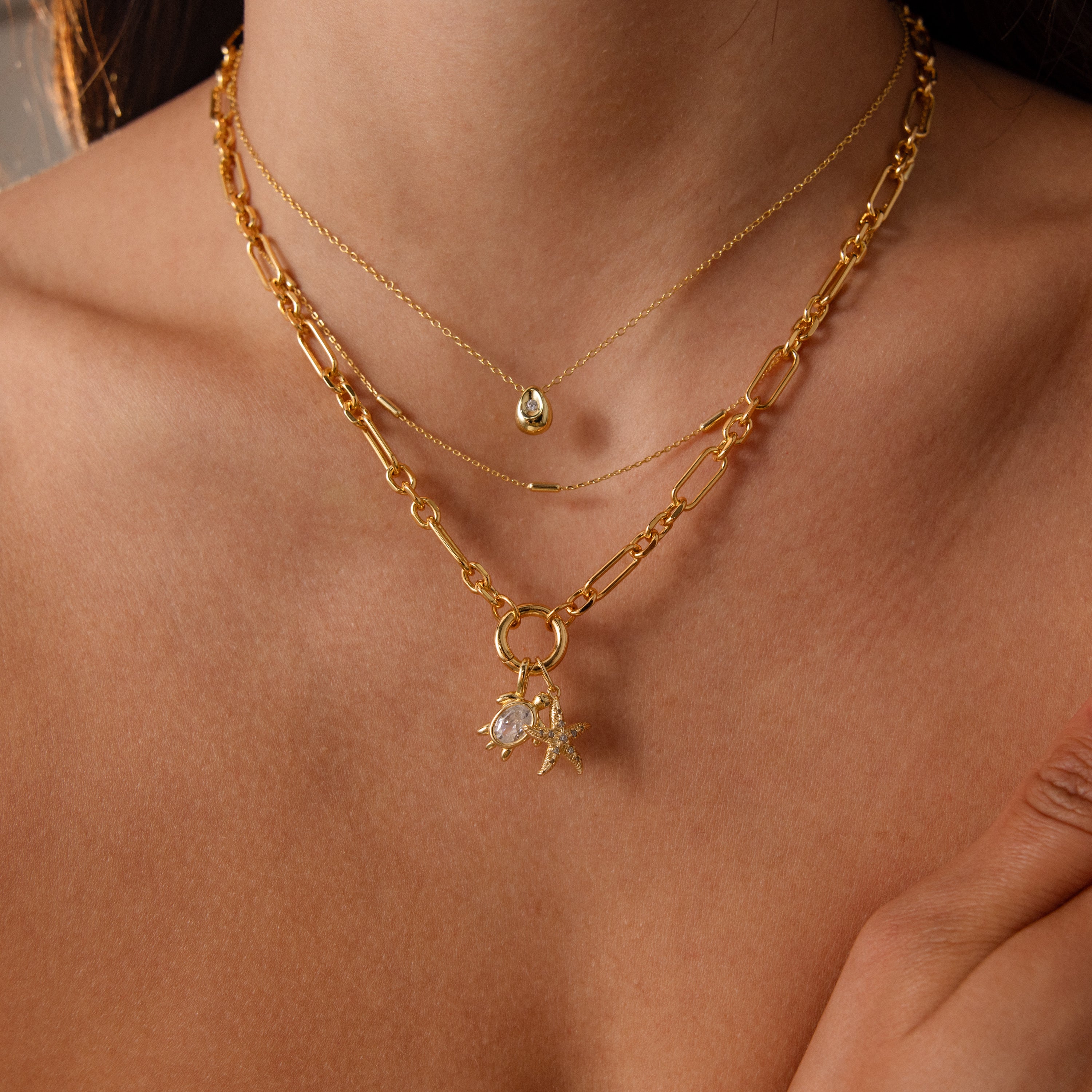 A woman wears layered gold necklaces against bare skin, including one with a Diamond Turtle Charm and another featuring a teardrop pendant.