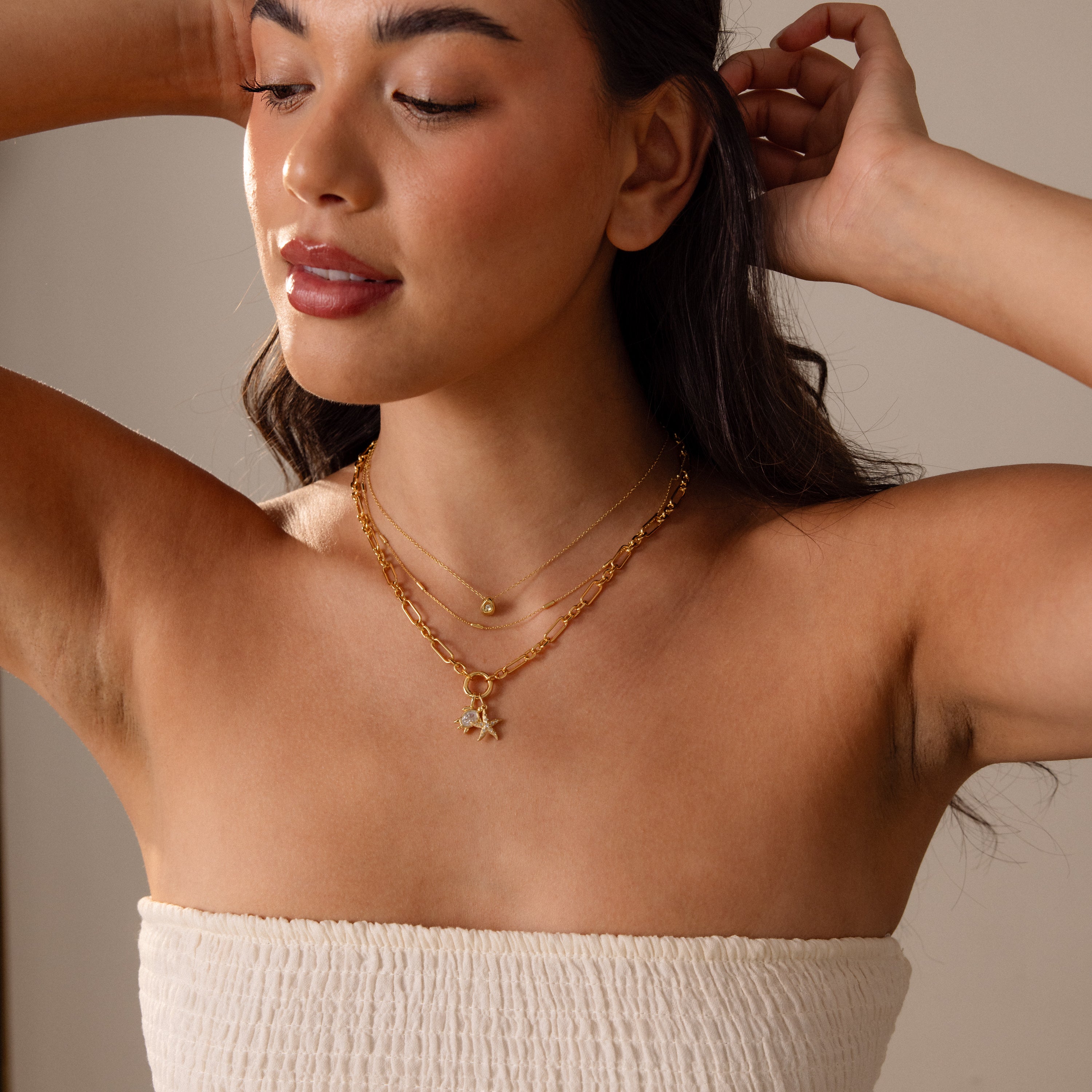 Woman in a white strapless top layers gold necklaces, featuring the Pave Starfish Charm in 18K Gold, and gazes to the side with one arm raised, exuding effortless coastal style.