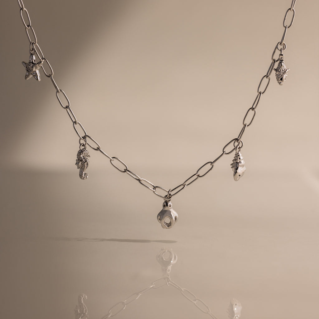 The Ocean Charm Necklace in Silver features sea-themed charms—shells and a starfish—for a laidback seaside look, shown on a beige background.