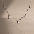 The Ocean Charm Necklace in Silver features sea-themed charms—shells and a starfish—for a laidback seaside look, shown on a beige background.