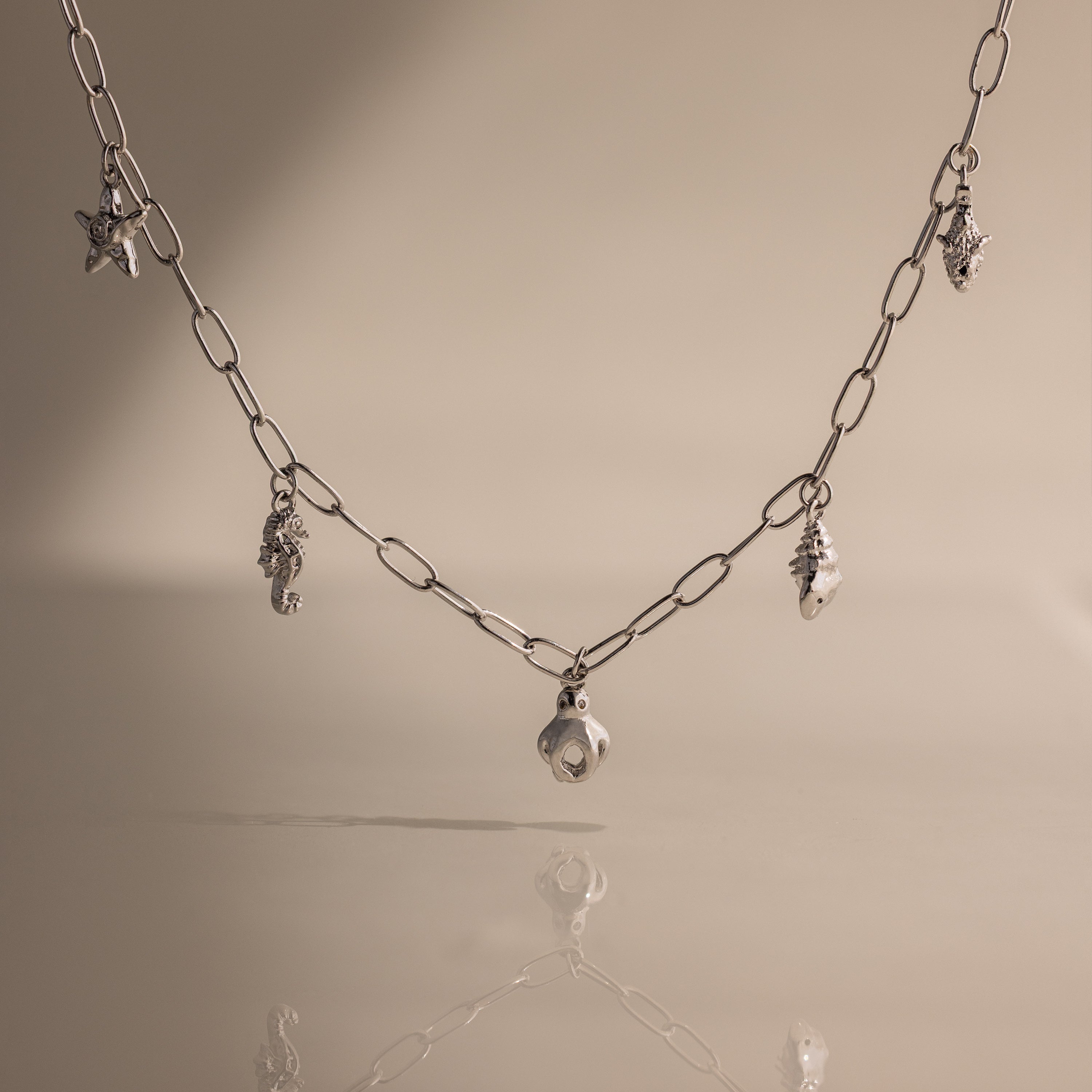 The Ocean Charm Necklace in Silver features sea-themed charms—shells and a starfish—for a laidback seaside look, shown on a beige background.