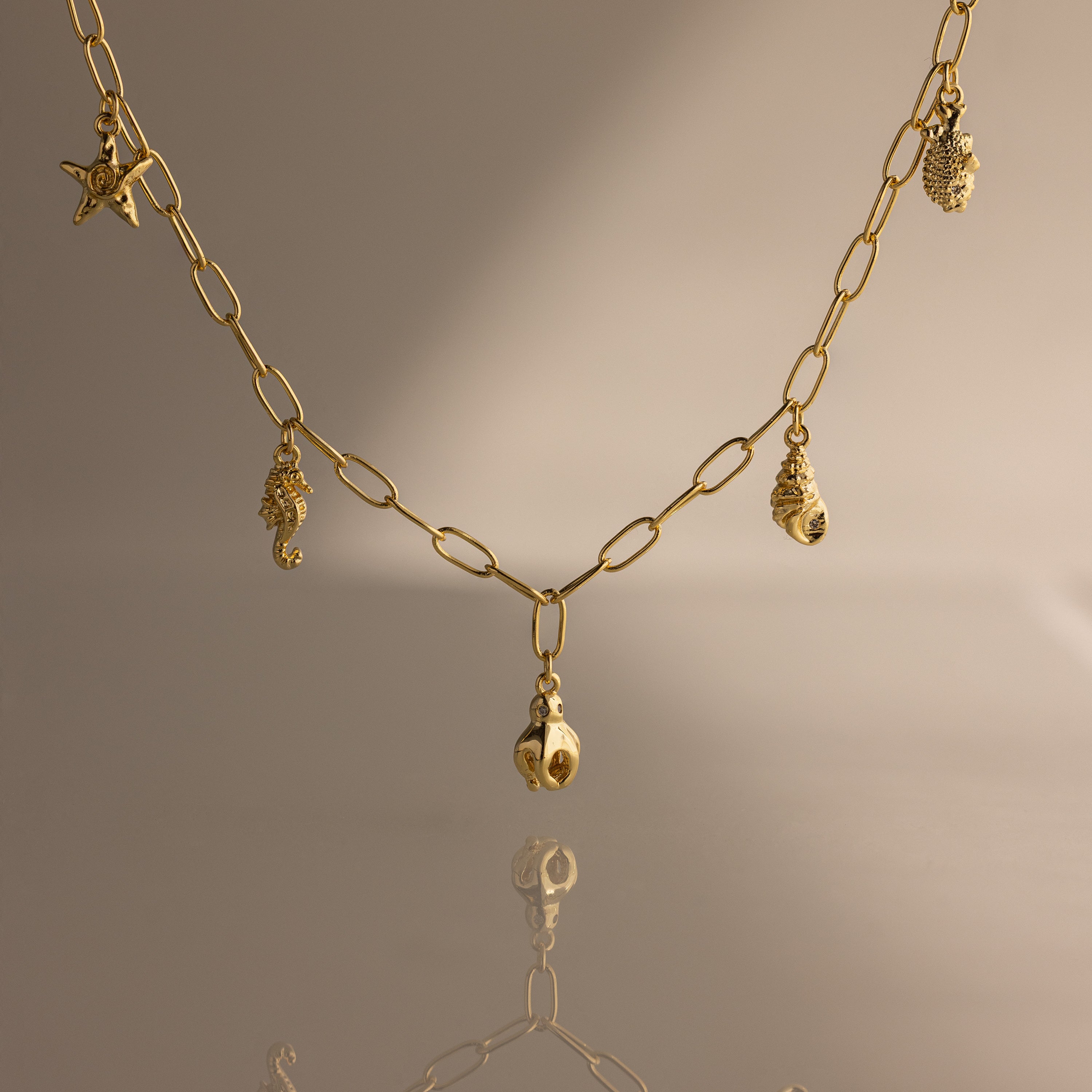 The Ocean Charm Necklace in 18K Gold features sea-themed charms—starfish, seahorse, shells, and pineapple—making it the perfect summer accessory on any neutral background.