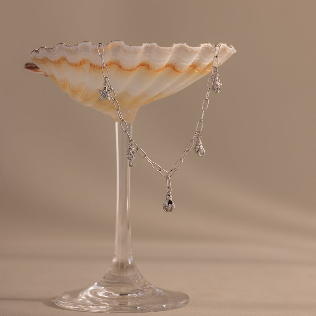 The Ocean Charm Necklace, in silver with seaside charms, is draped over a scallop shell and displayed on a glass stand against a beige background.