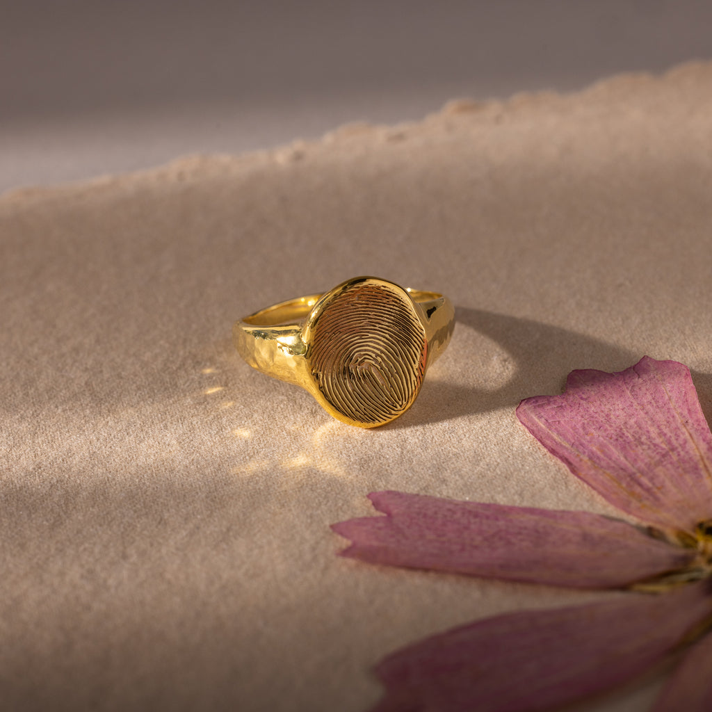 The Organic Fingerprint Signet Ring, featuring a unique design, rests on textured paper beside a dried pink flower—a perfect piece of personalized jewelry and a timeless keepsake.