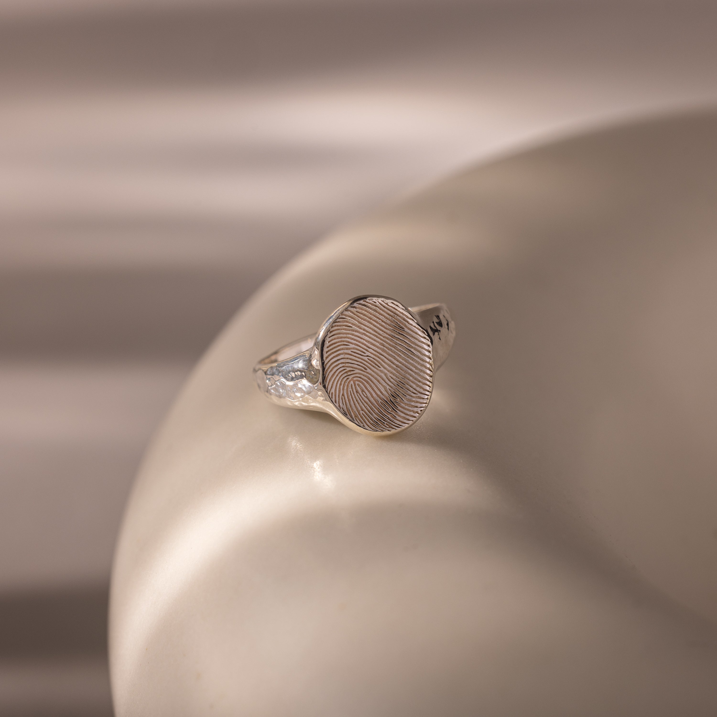 Organic Fingerprint Signet Ring: This handcrafted silver ring features a unique fingerprint design, displayed on a smooth beige surface with soft lighting for an elegant, personalized keepsake.