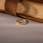 The Hand & Footprint Signet Ring in 18K Gold, featuring an engraved design, rests on a gray surface beside a closed leather-bound book.