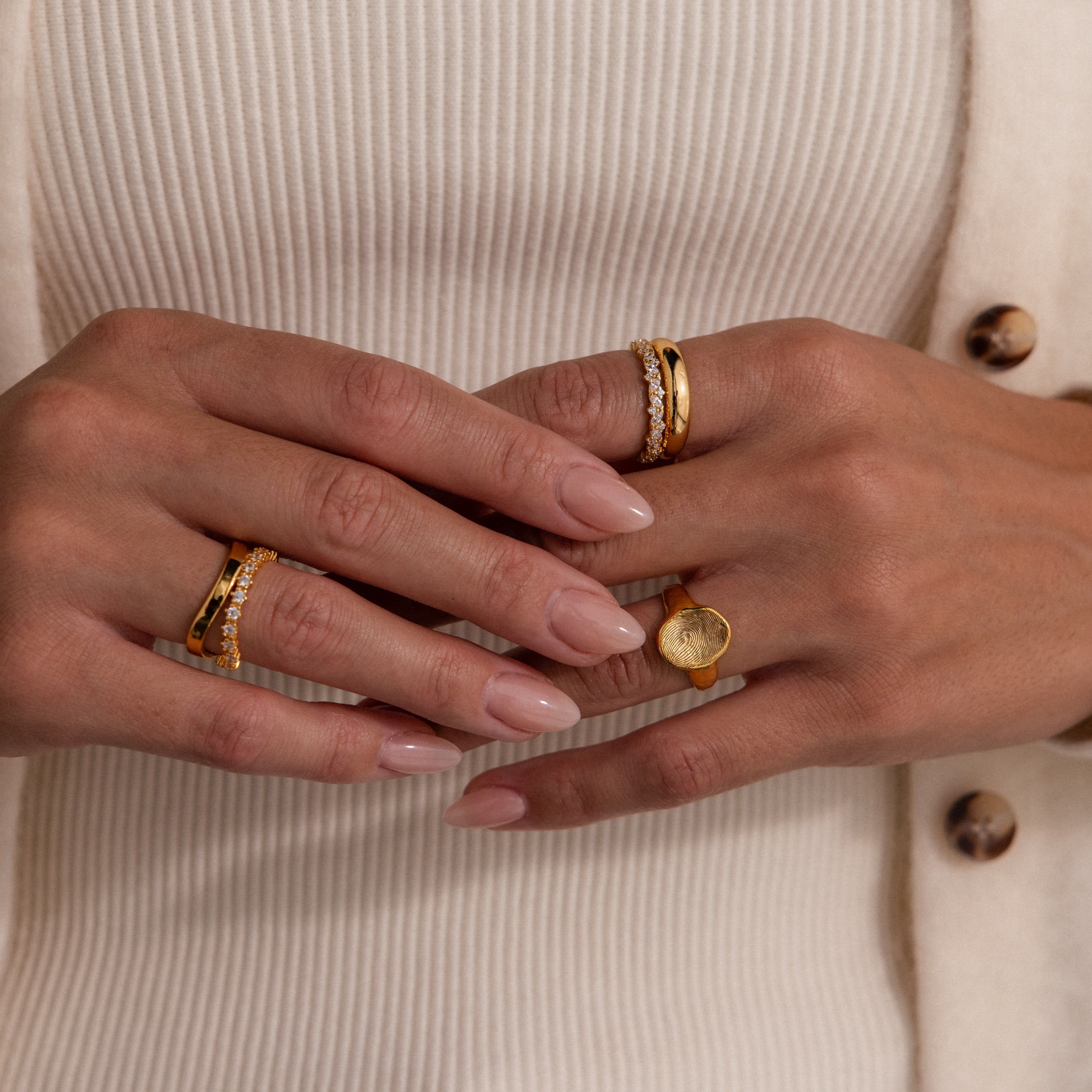 Close-up of manicured hands adorned with gold rings, including the Organic Fingerprint Signet Ring, against a cream ribbed top and buttoned sweater—a chic take on personalized jewelry.