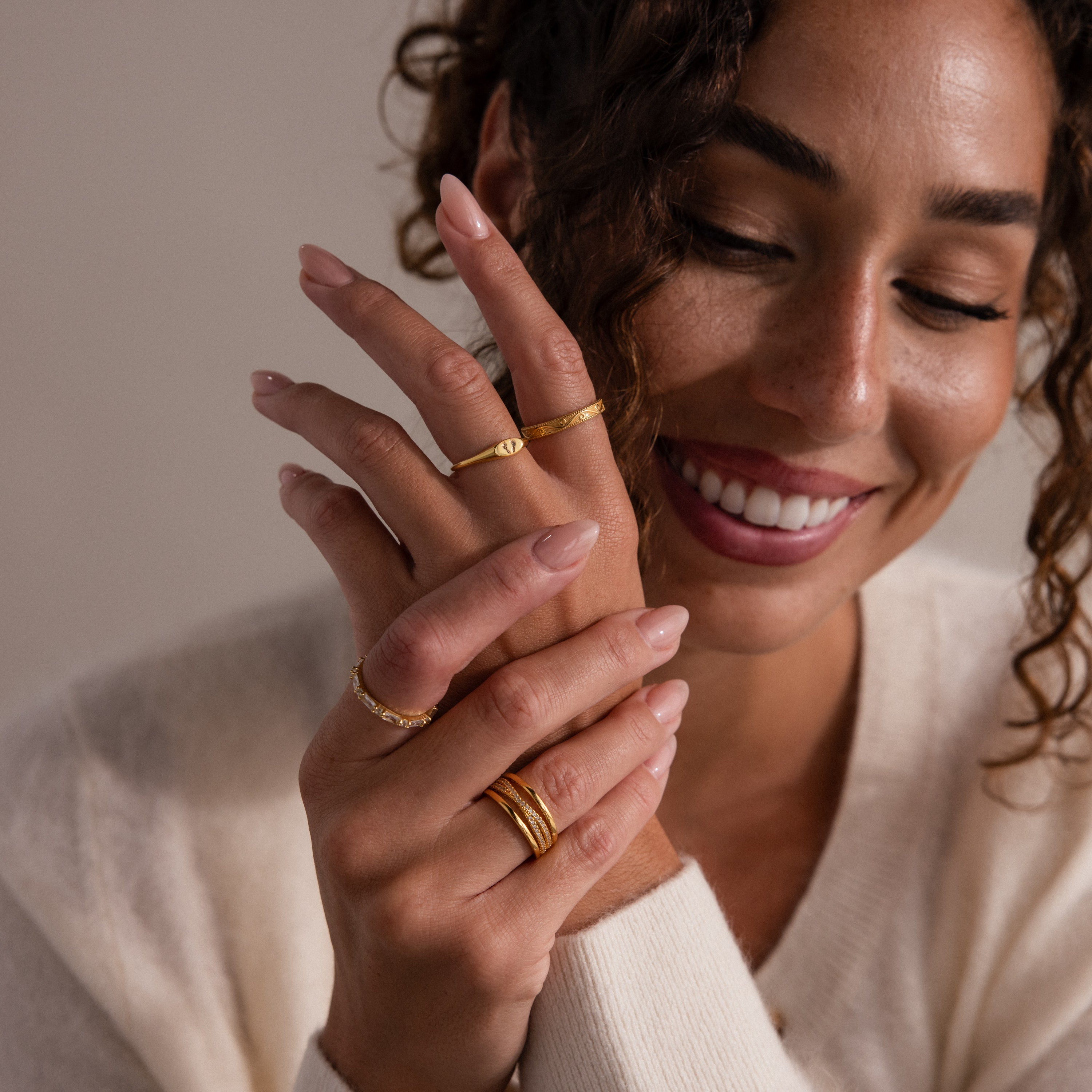 A smiling woman with curly hair wears a cream sweater and showcases the Hand & Footprint Signet Ring in Rose Gold, along with other gold rings, on her hands.