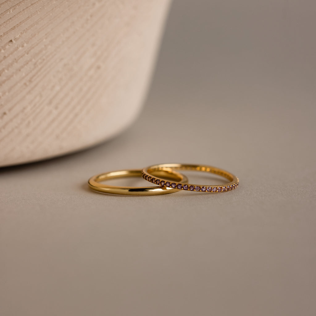 Close-up of a delicate gold band ring set with a continuous row of sparkling alexandrite gemstones, showcasing their soft violet-lavender hue. The ring rests on a sculptural, ribbed ceramic surface in a warm neutral tone, emphasizing the contrast between the luminous gold finish and the subtle shimmer of the birthstones.