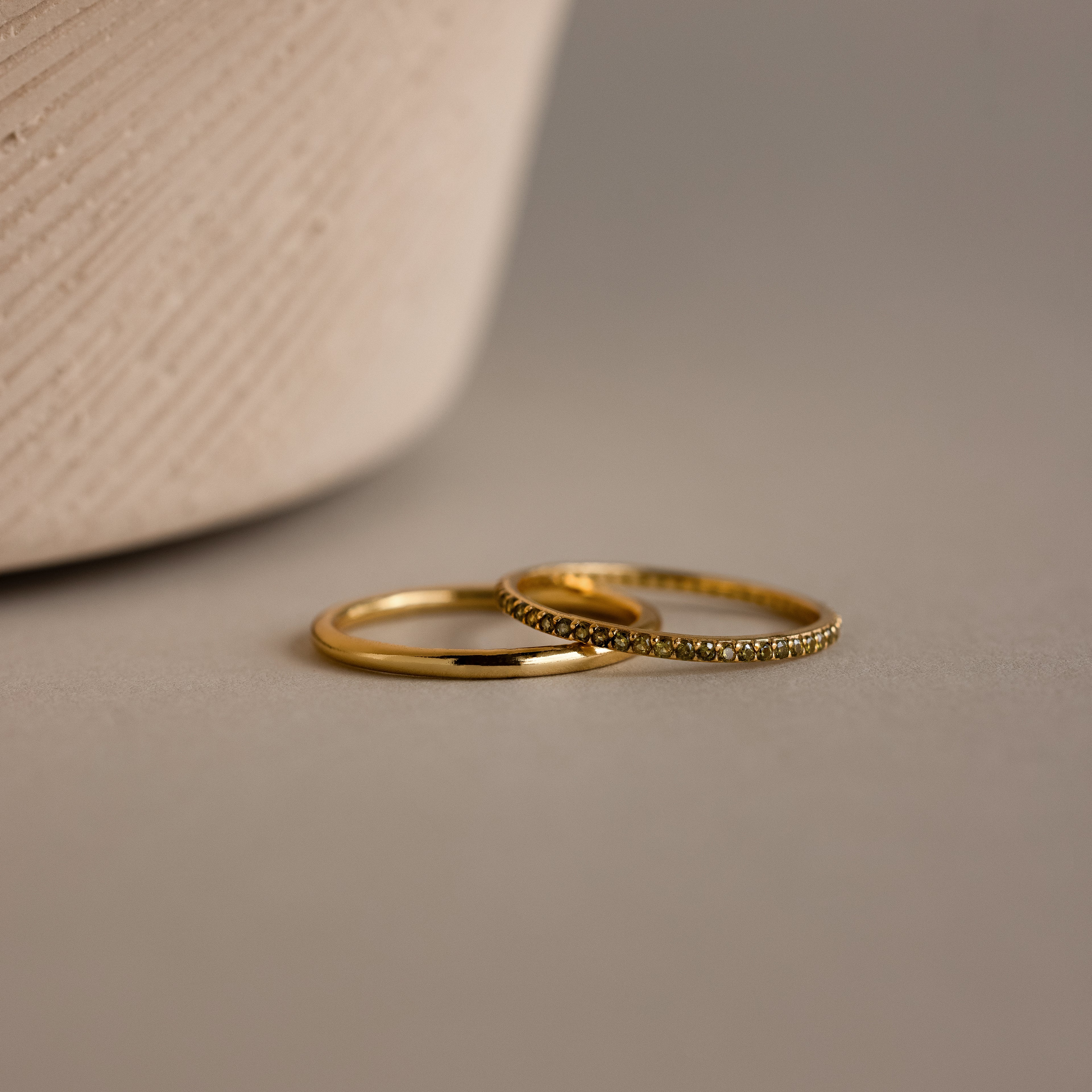 The Peridot Tennis Ring Set, featuring one simple gold band and one gold eternity ring with small peridot stones, is displayed on a neutral surface beside a textured object.