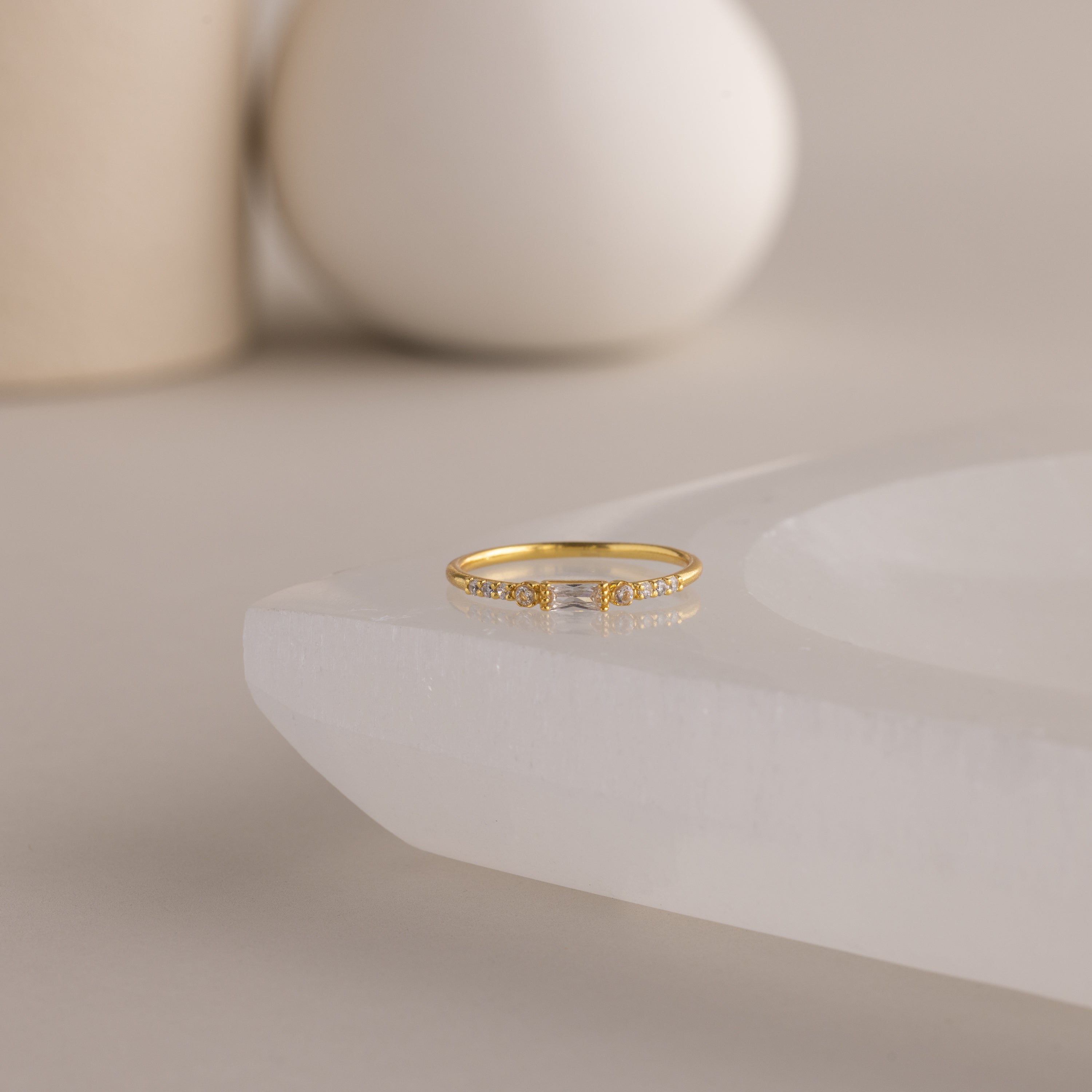 A single dainty gold birthstone ring with a rectangular diamond gemstone and small white accent stones rests on a curved white stone surface with minimalist background decor.
