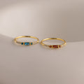 Two delicate gold birthstone rings side by side on a white stone slab—one with a garnet baguette gemstone, the other with a vibrant blue zircon baguette stone—each accented with petite clear stones along the band.