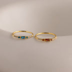 Two delicate gold birthstone rings side by side on a white stone slab—one with a garnet baguette gemstone, the other with a vibrant blue zircon baguette stone—each accented with petite clear stones along the band.