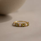 The Peridot & Pearl Lace Ring features a gold band with alternating green peridots and pearls, showcased on a neutral surface against a soft background.