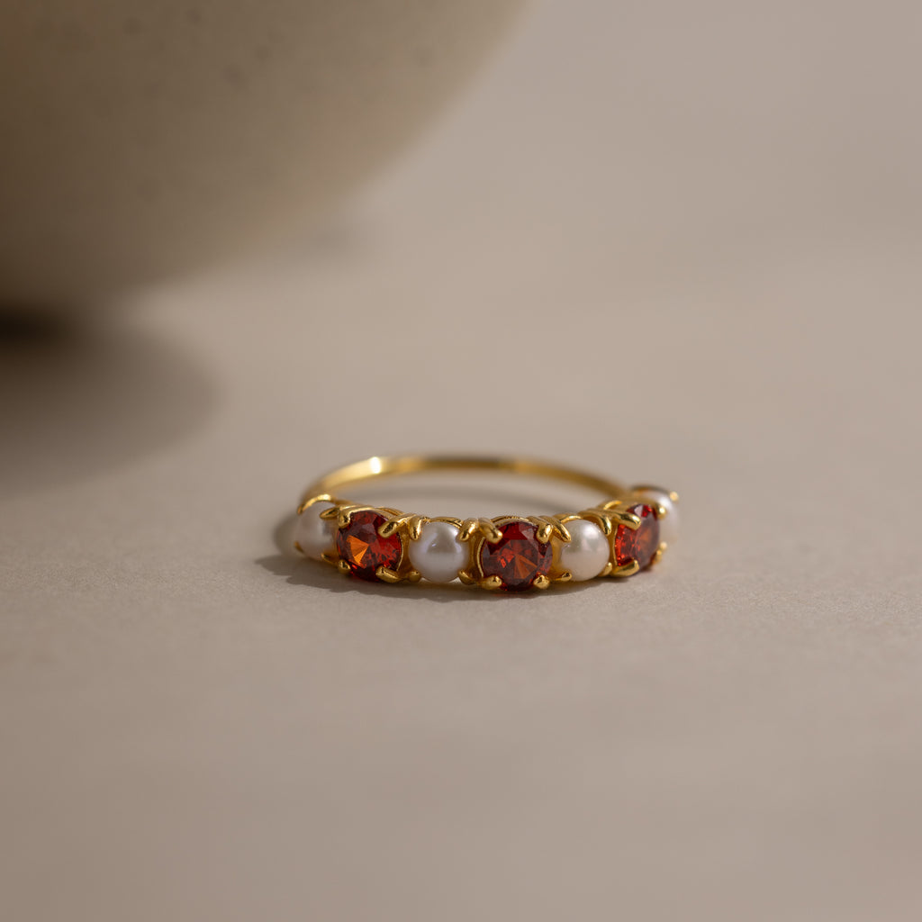The Birthstone & Pearl Lace Ring in Garnet (January) features a gold band with alternating red gemstones and white pearls, displayed on a neutral background.
