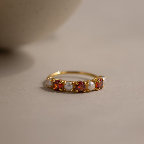 The Birthstone & Pearl Lace Ring in Garnet (January) features a gold band with alternating red gemstones and white pearls, displayed on a neutral background.