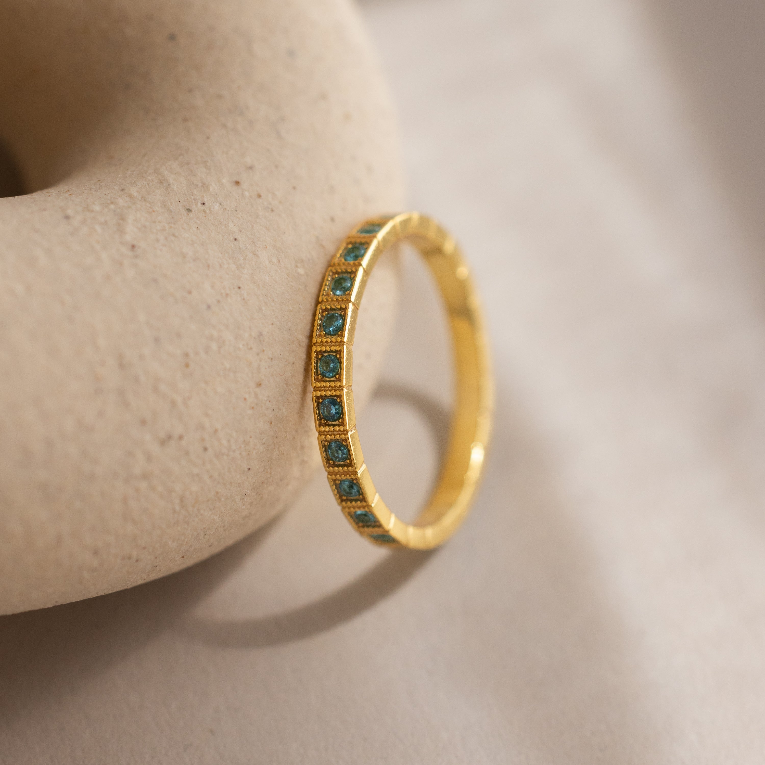 The Art Deco Aquamarine Eternity Ring features a vintage-inspired gold band with small green stones, displayed on a textured beige surface for a look of timeless elegance.
