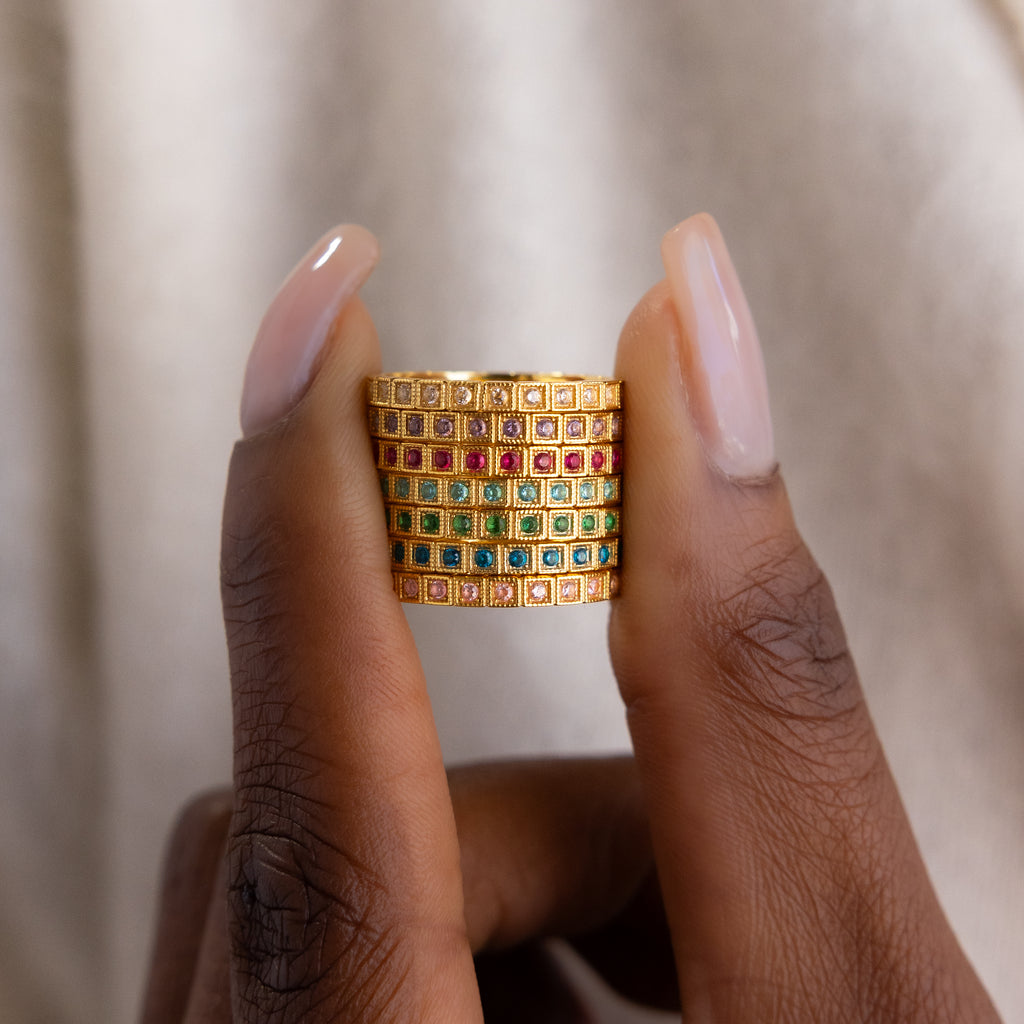A hand holding an Art Deco Diamond Eternity Ring with a wide gold band set in a grid pattern with red, green, and purple square gemstones.