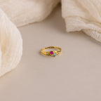 Toi et Moi Interlocking Name Ring in 18K Gold, featuring three gemstones and engraved words, displayed on a neutral surface with soft fabric accents.
