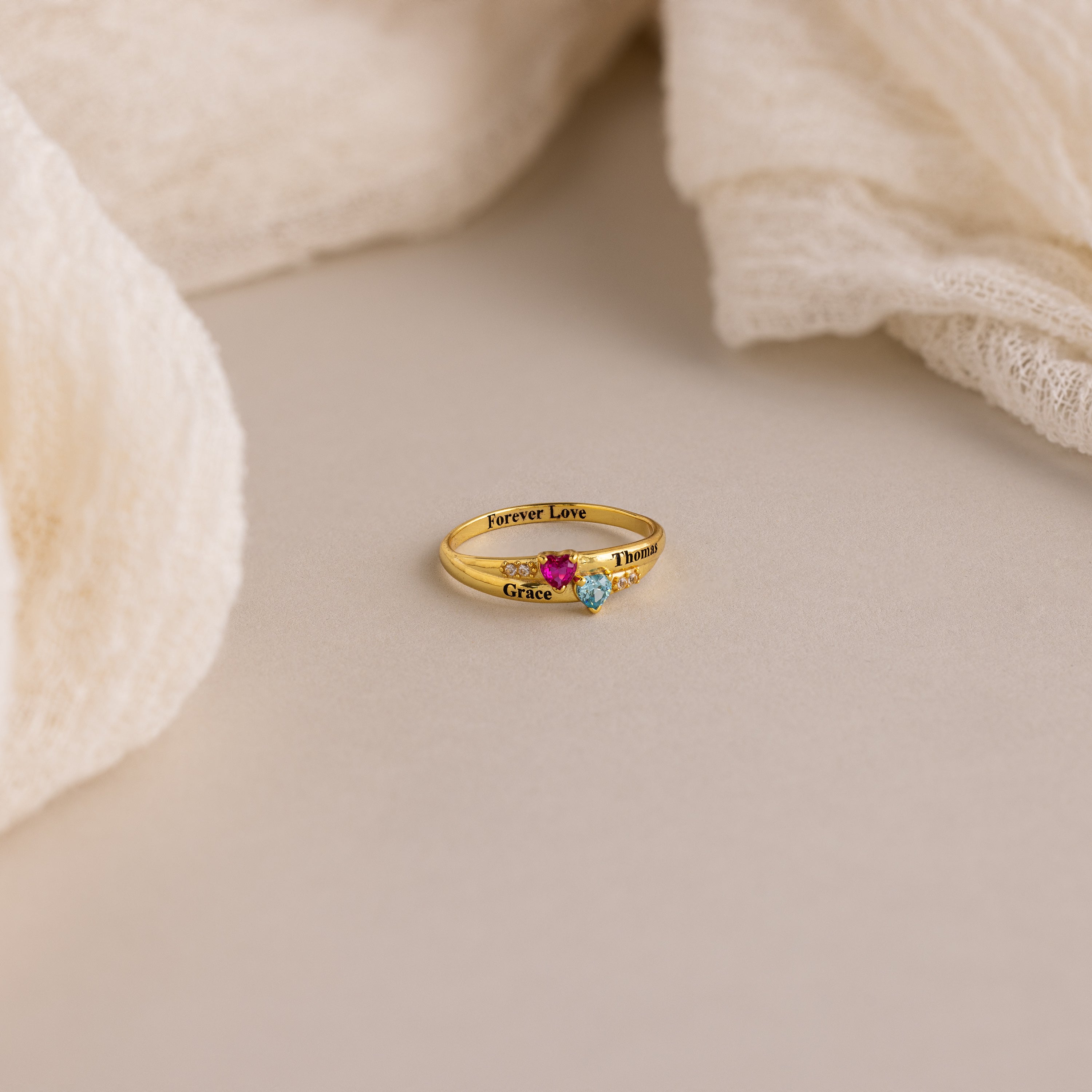 Toi et Moi Interlocking Name Ring in 18K Gold, featuring three gemstones and engraved words, displayed on a neutral surface with soft fabric accents.