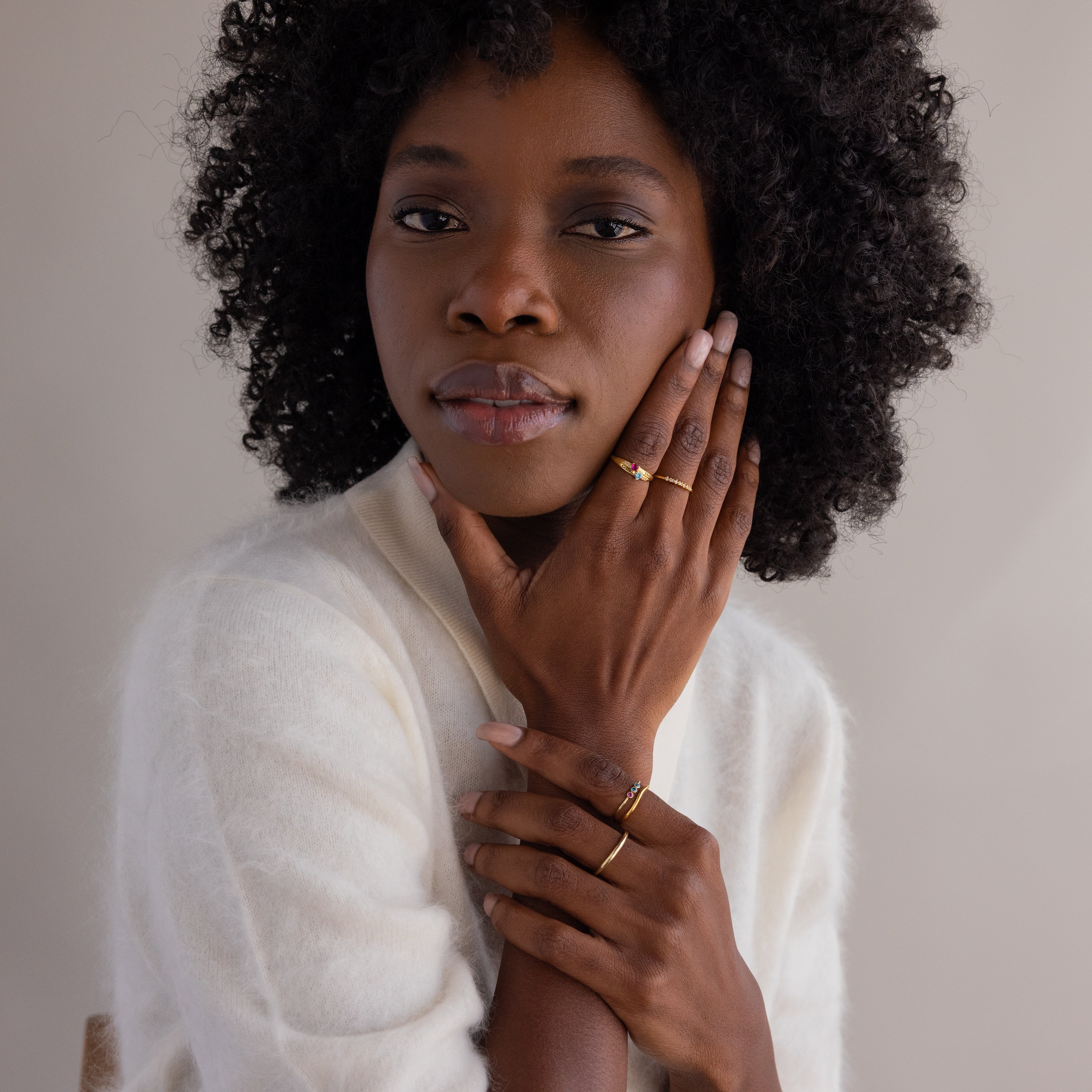 A woman with curly hair in a white sweater touches her face, displaying the Toi et Moi Interlocking Name Ring in 18K Gold as she gazes at the camera.