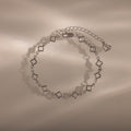 The Clover Station Bracelet in Silver features delicate clover-shaped links and a heart charm, elegantly displayed on a beige surface.