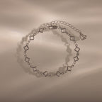 The Clover Station Bracelet in Silver features delicate clover-shaped links and a heart charm, elegantly displayed on a beige surface.